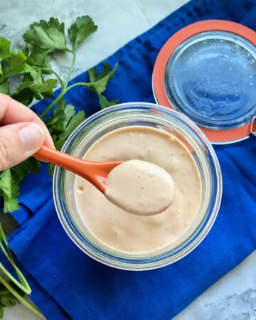 Top view of a female hand holding a small orange spoon dipped in a jar of sauce.