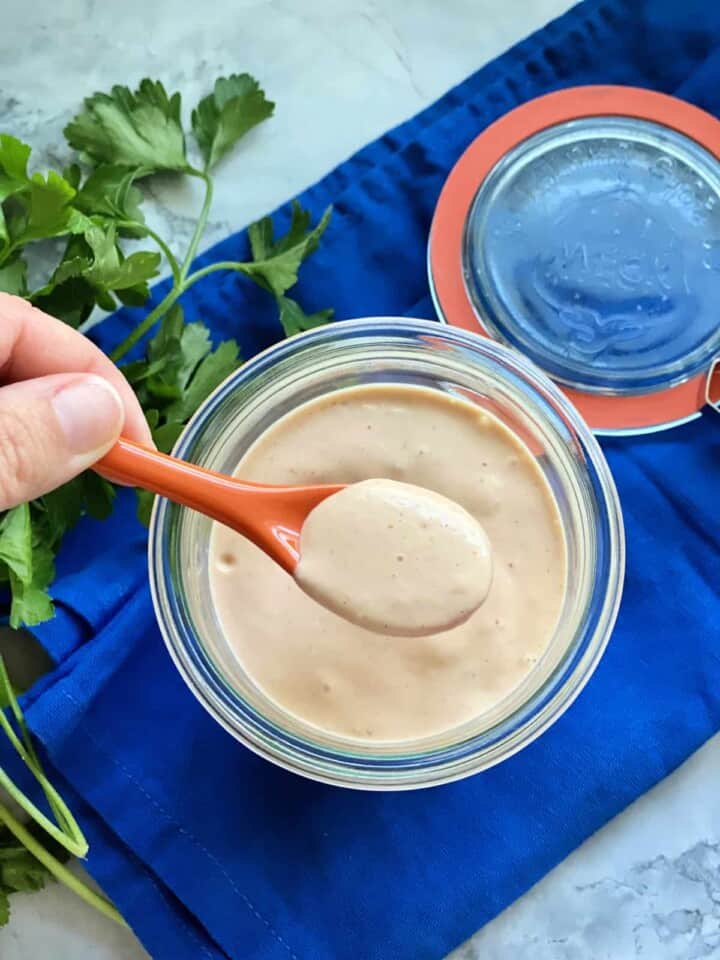 Top view of a female hand holding a small orange spoon dipped in a jar of sauce.