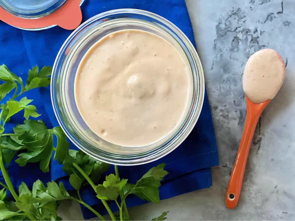 Top view of a glass jar filled with a light color sauce and an orange spoon filled with the sauce layin next to it on the counter.