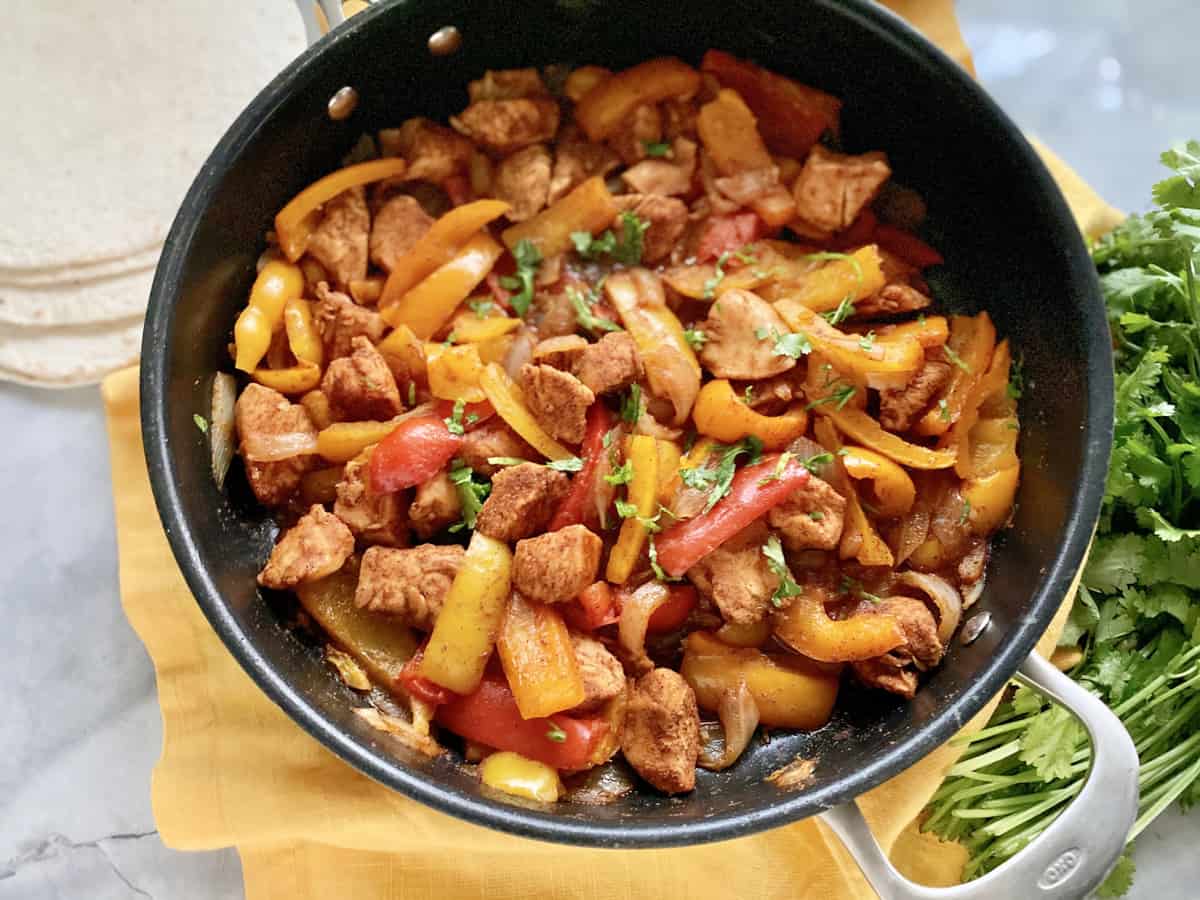 Top view of a skillet full of bell peppers and chicken on a yellow cloth with cilantro next to it.