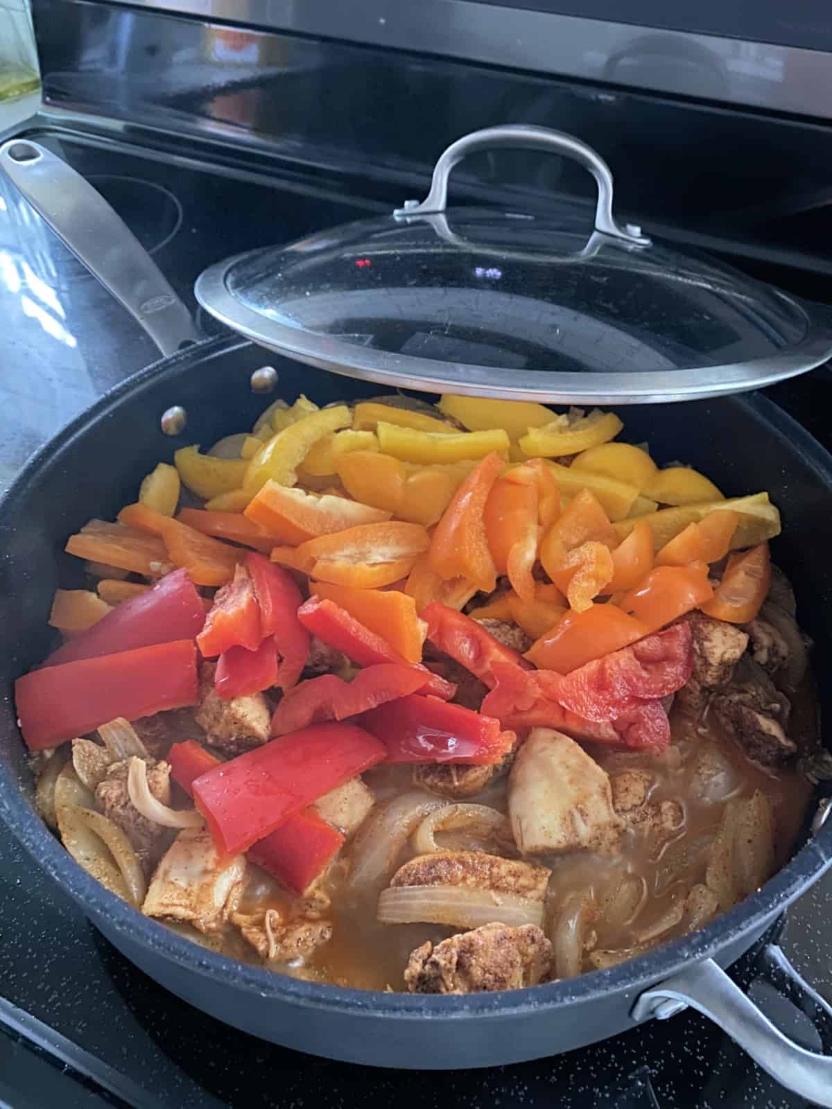 Large skillet with lid draped off the side with yellow, orange, and red bell peppers and cooked chicken.