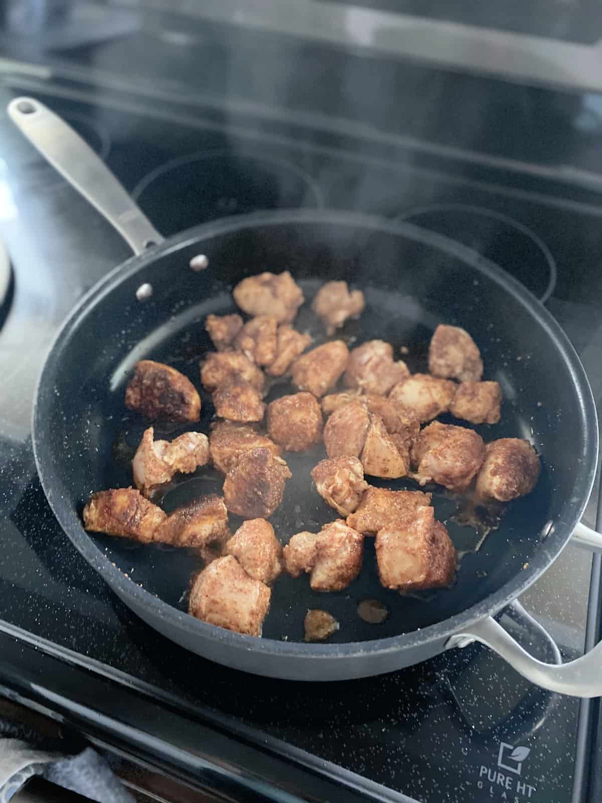 Skillet on a glass stove top cooking cubed chicken breast with seasoning.