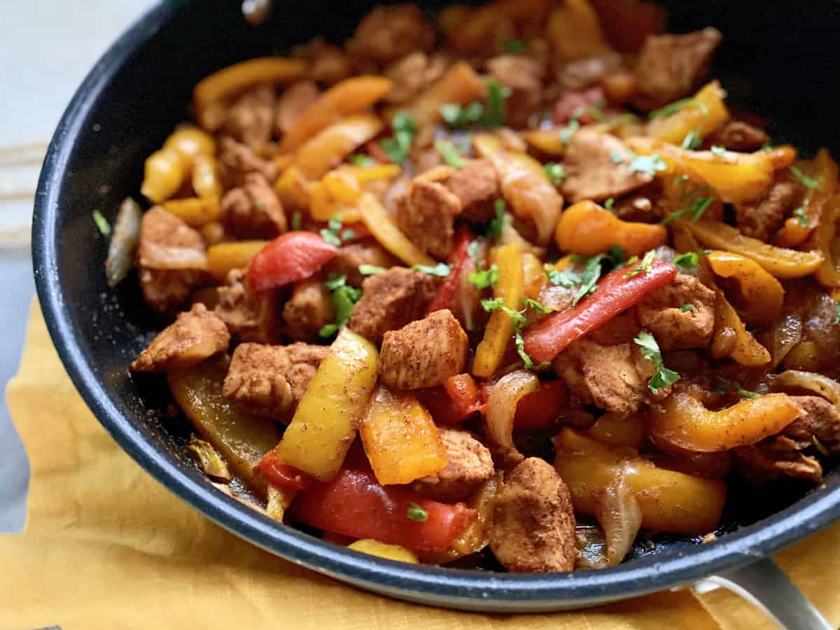 Skillet with bell peppers, onions, and chicken resting on a yellow cloth.