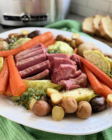 White platter filled with corned beef, cabbage, carrots, and potatoes with slow cooker in background.