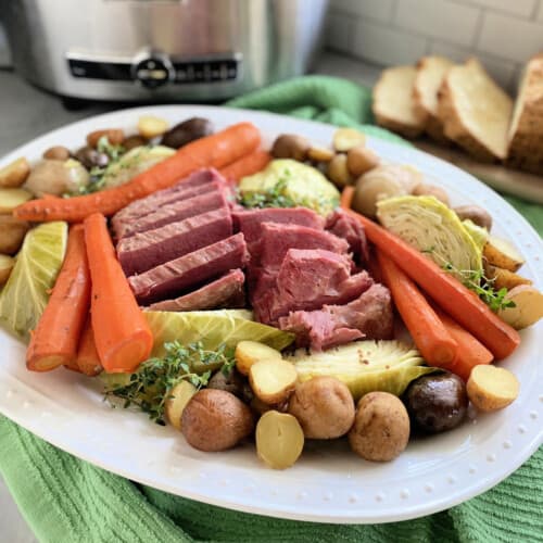 White platter filled with corned beef, cabbage, carrots, and potatoes with slow cooker in background.
