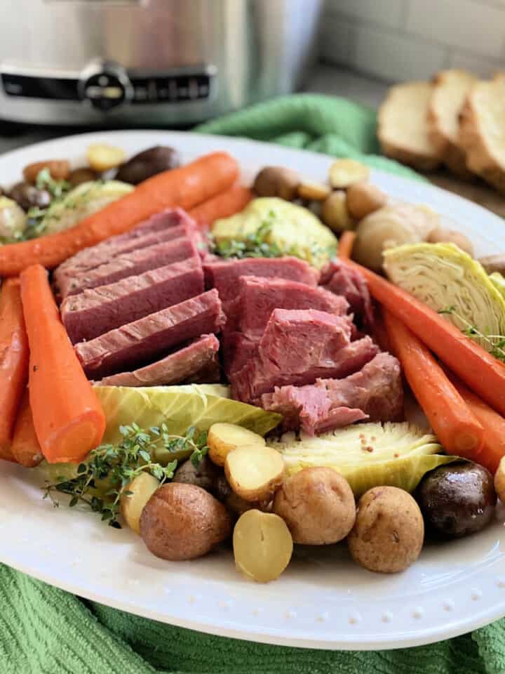 White platter filled with corned beef, cabbage, carrots, and potatoes with slow cooker in background.
