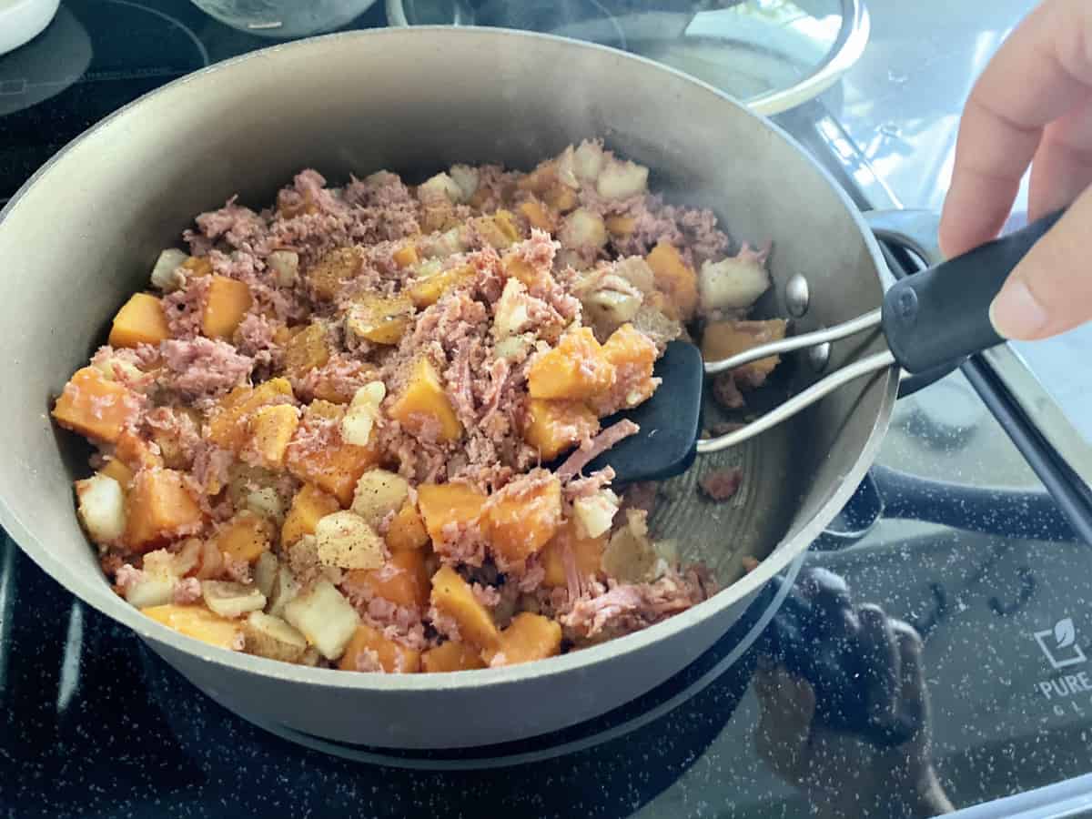 Female hand flipping Sweet Potato Corned Beef Hash in a frying pan on a glasstop cook top.