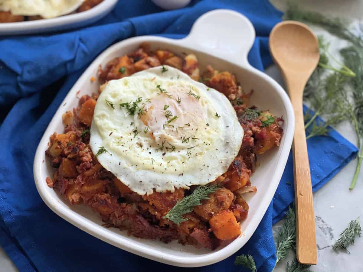 White square plate filled with Sweet Potato Corned Beef Hash with a fried egg on top.