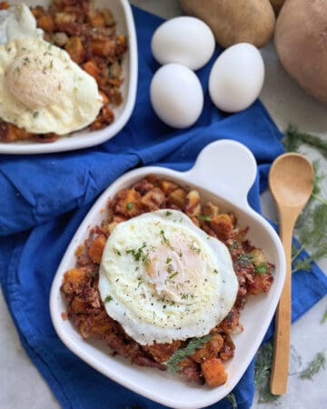 Top view of a white square plate filled with corned beef hash and fried eggs with a wooden spoon and eggs next to it.