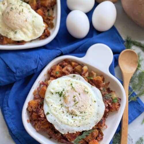 Top view of a white square plate filled with corned beef hash and fried eggs with a wooden spoon and eggs next to it.