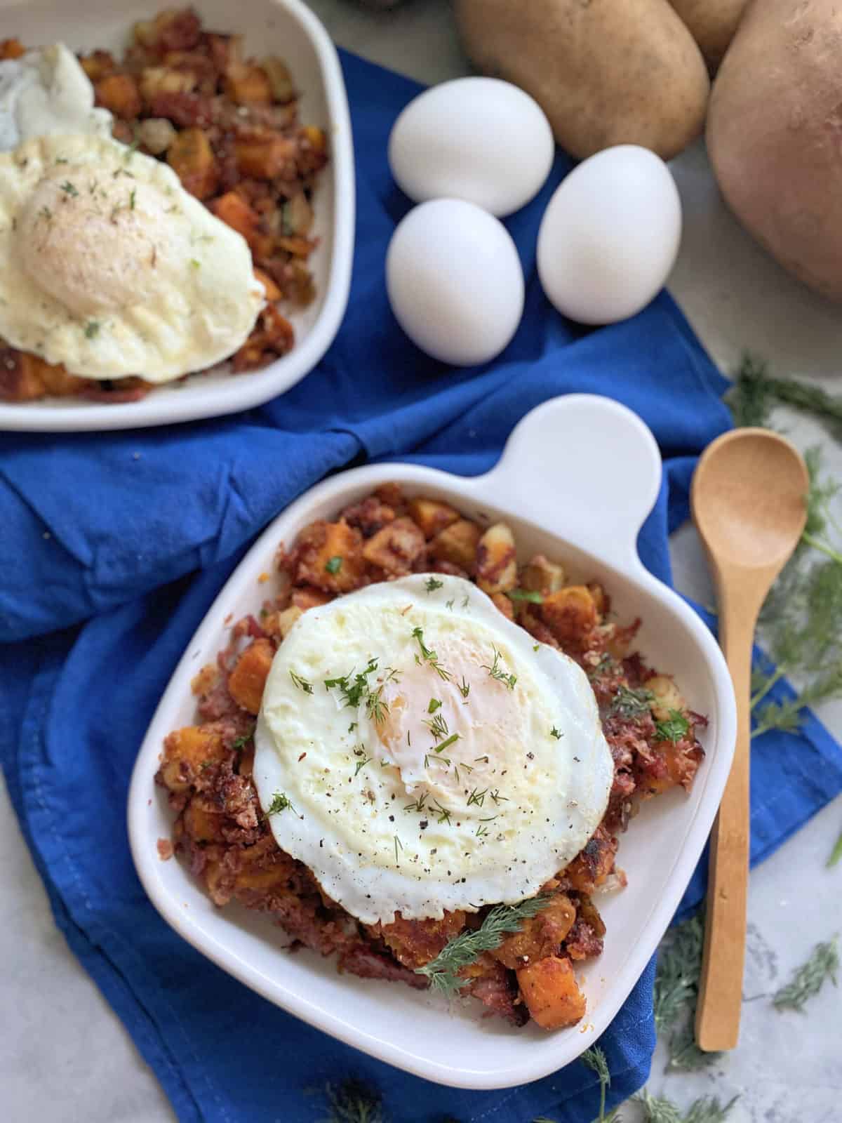 Top view of a white square plate filled with corned beef hash and fried eggs with a wooden spoon and eggs next to it.