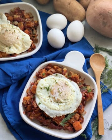 Two square white plates with corned beef hash, fried eggs, and dill on top.