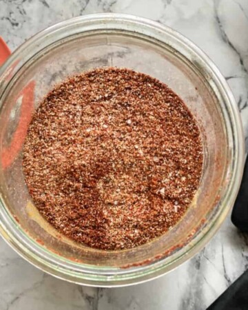 Top view of a glass filled with seasoning and measuring spoons laying on marble counter next to glass jar.