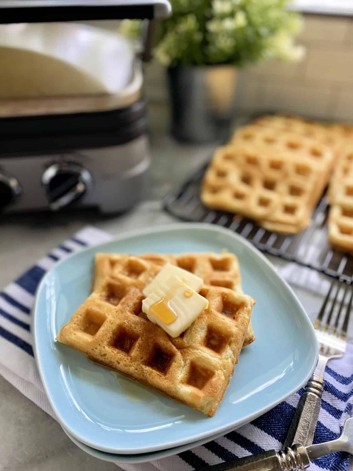Blue plate with two rectangular waffles with butter and syrup on them with waffles in background.