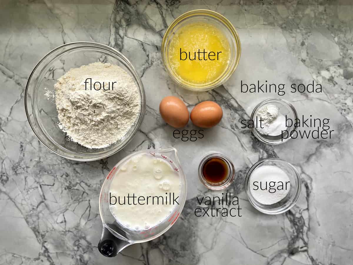 Ingredients on marble countertop: flour, buttermilk, butter, baking soda, salt, baking powder, vanilla extract, sugar, and eggs.