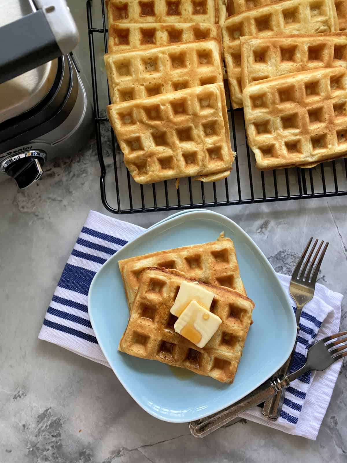 Top view of a blue plate filled with two waffles iwth butter and syrup with waffles and iron around it.