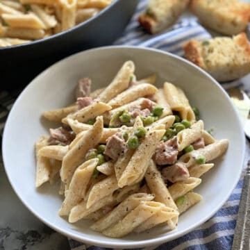 White bowl filled with creamy penne pasta, ham, and peas with a skillet and bread in the background.
