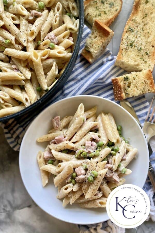 Top view of a bowl of pasta with a skillet with bread next to it and logo on right corner.