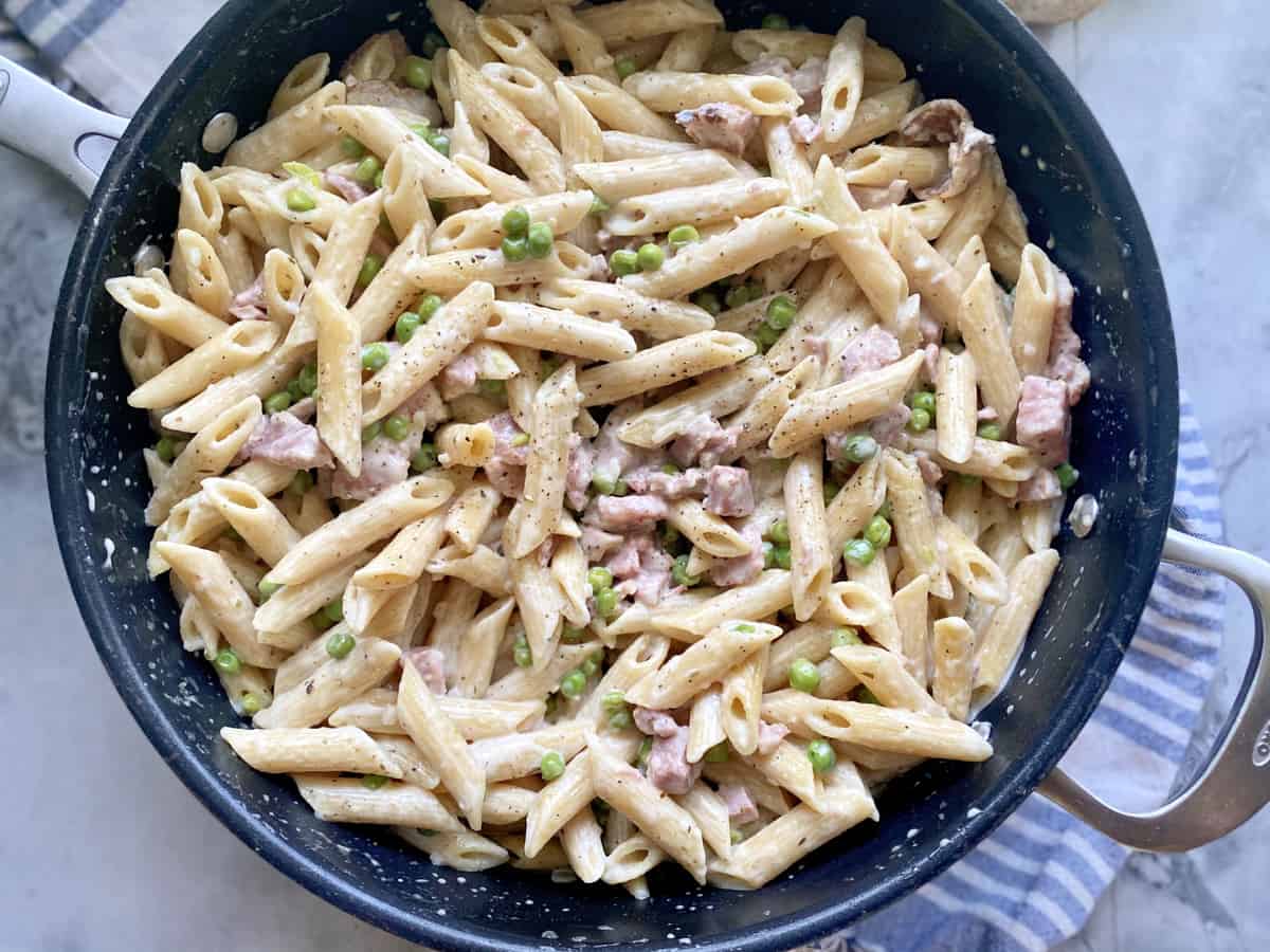 Top view of a large black skillet filled with creamy penne pasta, ham, and peas.