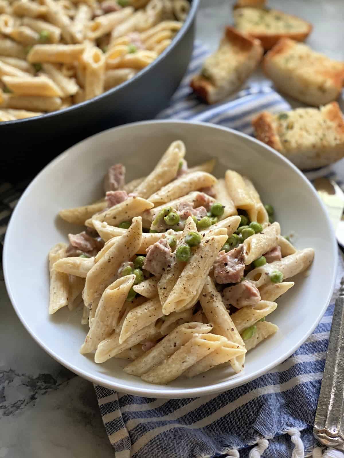 White bowl filled with creamy penne pasta, ham, and peas with a skillet and bread in the background.