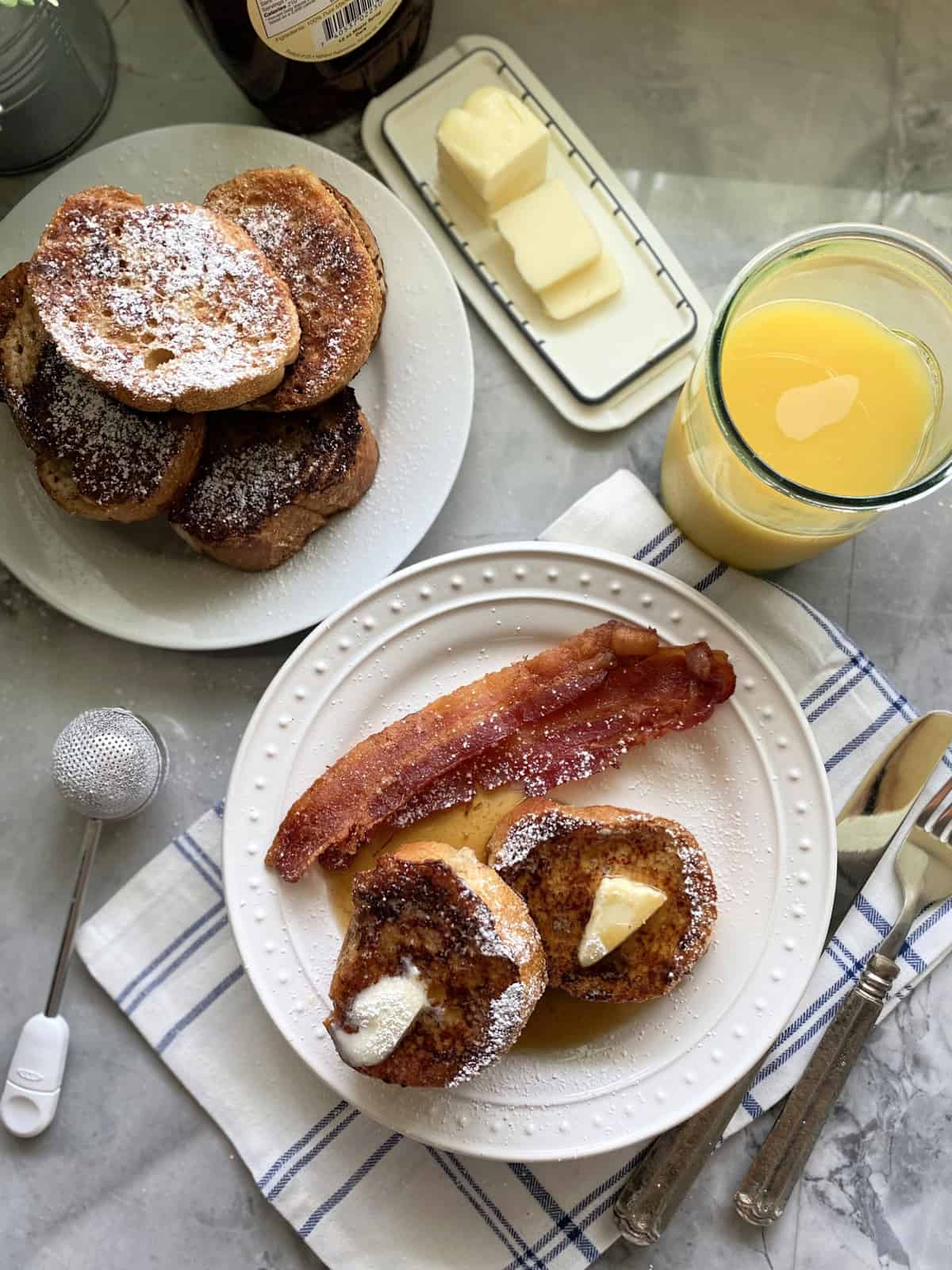 Top view of a stack of french toast and another plate filled with french toast, butter, syrup, and bacon.