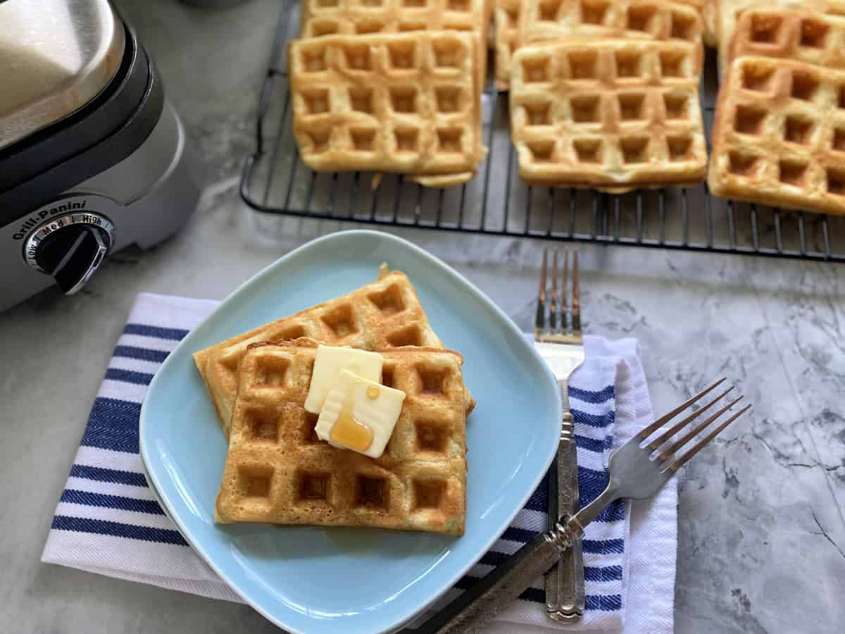 Top view of a blue plate with two waffles with wire rack full of waffles next to it.