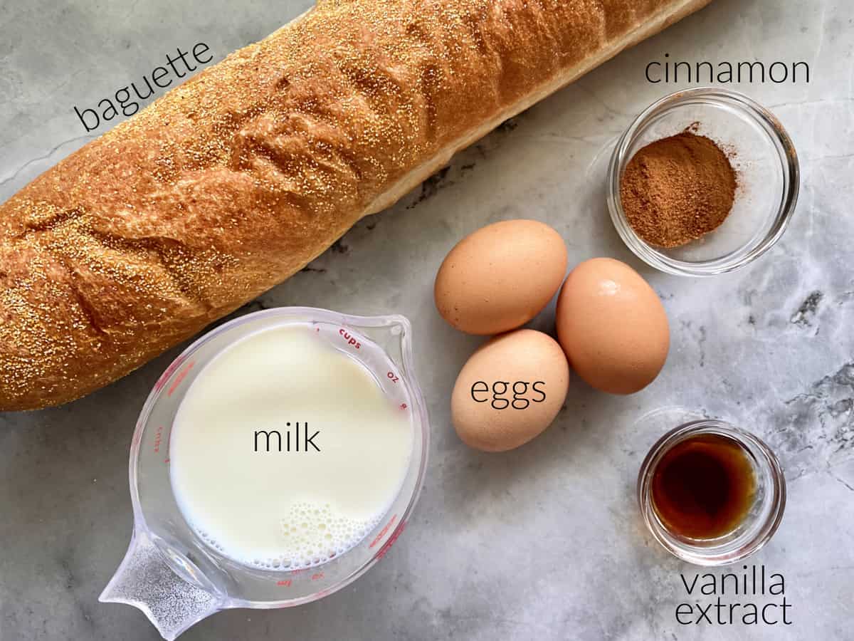 Ingredients on marble countertop: baguette, milk, eggs, cinnamon, vanilla extract.