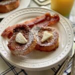 White plate filled with 2 slices of french toast with butter and syrup with bacon slices on white plate.