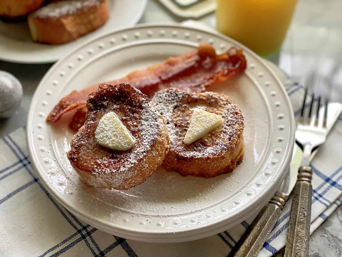 White plate filled with 2 slices of french toast with butter and syrup with bacon slices on white plate.