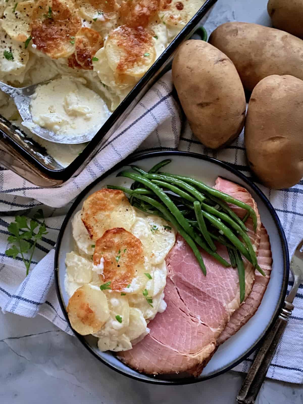 Top view of a plate filled with ham, creamy potatoes, and green beans with a baking dish filled with creamy potatoes next to it.