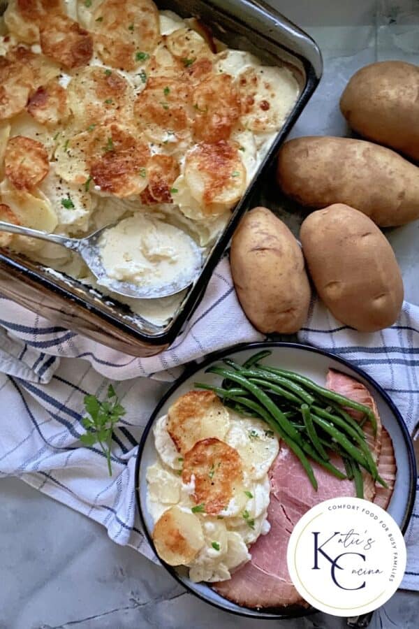 Top view of a baking dish filled with Gratin Dauphinois and a plate of ham, potatoes, and green beans.