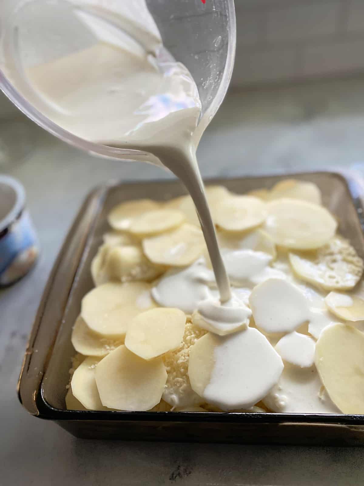 Measuring cup with milk pouring into a baking dish filled with sliced potatoes.