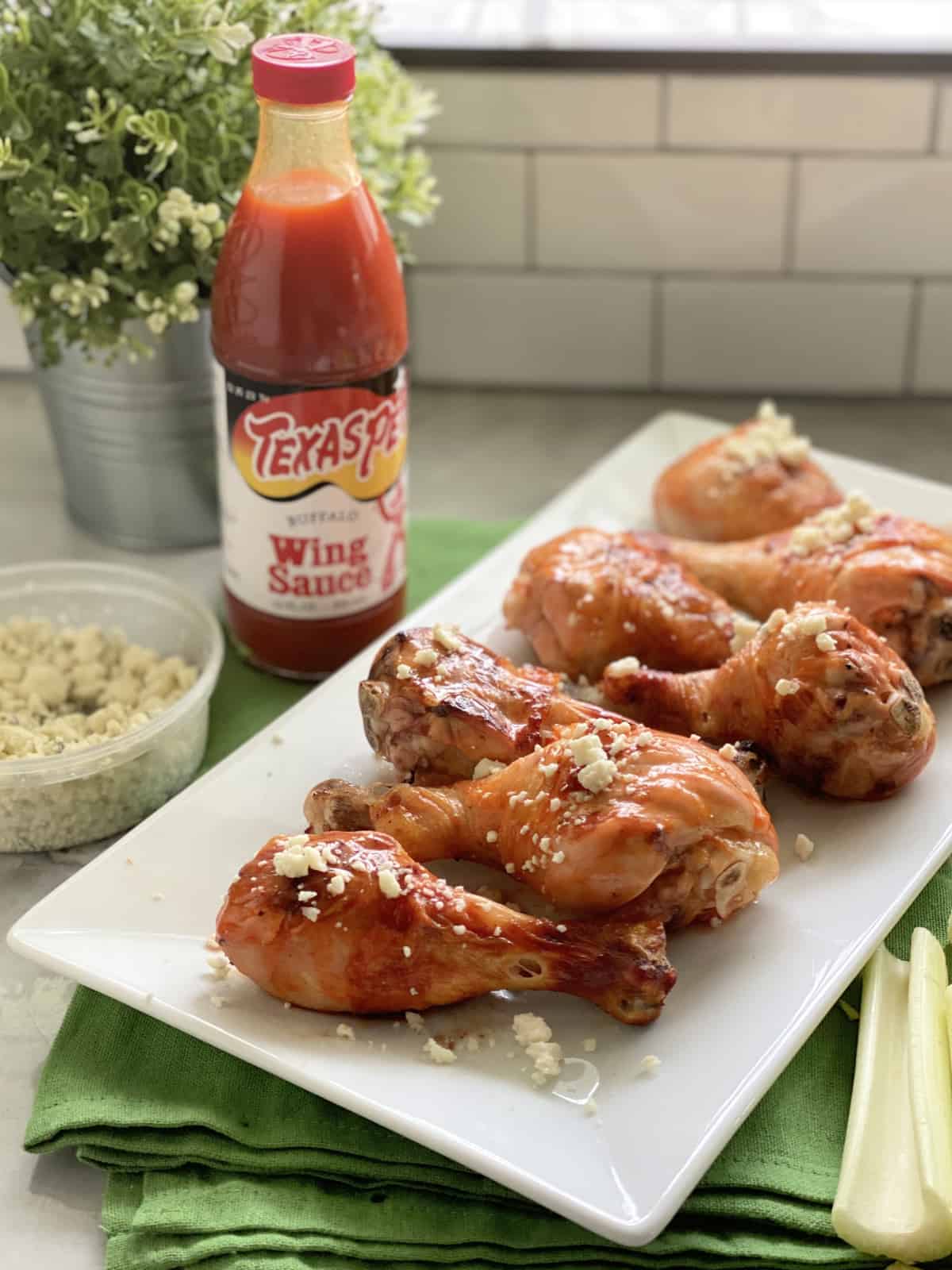 White platter filled with chicken drumsticks with buffalo sauce bottle in the background.