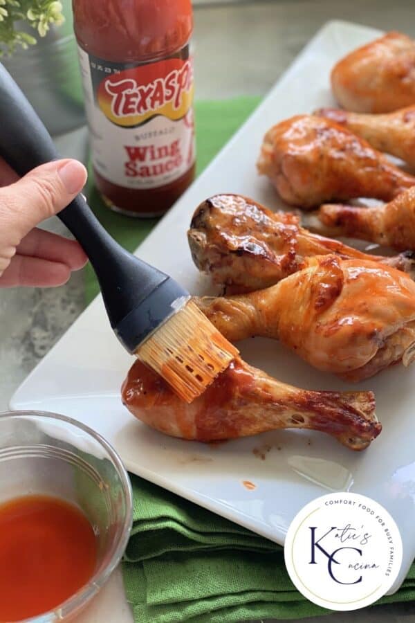 Female hand brushing buffalo sauce on chicken drumsticks with logo on the right corner.