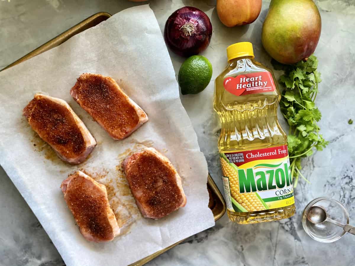 Baking sheet lined with white parchment paper with raw seasoned pork chops with fruit and a bottle of Mazola® Corn Oil next to it.