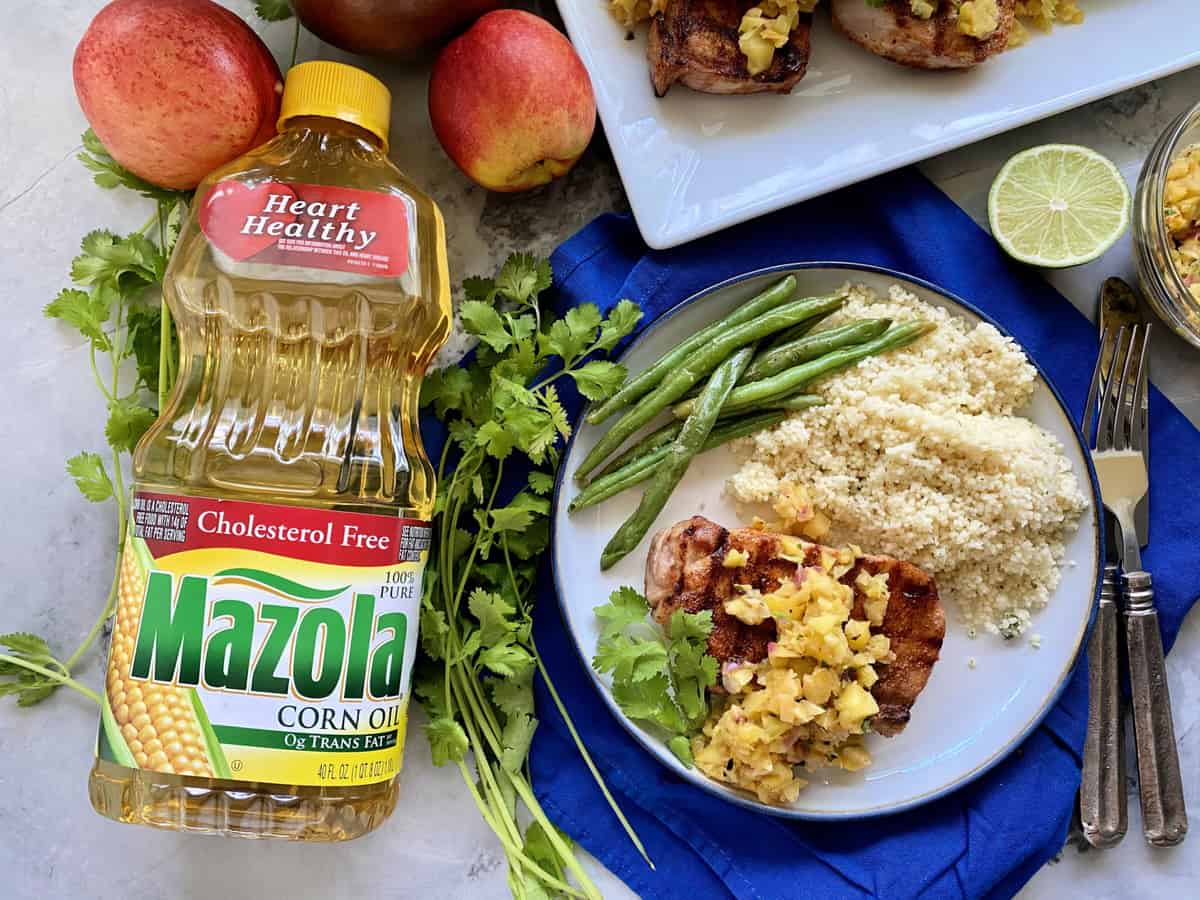 Top photo of a bottle of Mazola® Corn Oil with a plate of grilled pork chops, fruit salsa, green beans, and couscous on a blue cloth.