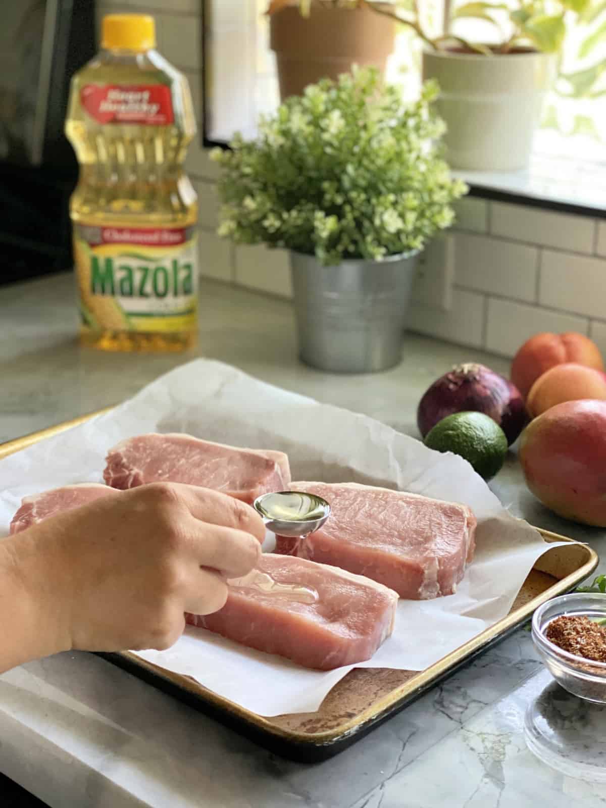 Female hand pouring a tablespoon of Mazola® Corn Oil over raw pork chops on a baking sheet.