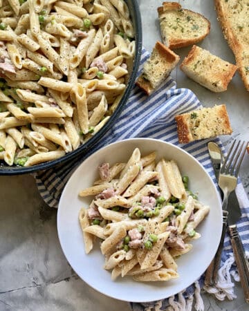 Top view of a skillet and white bowl filled with penne pasta, ham, and green peas.