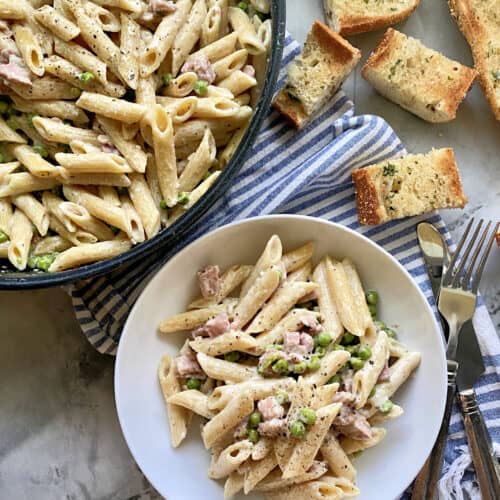 Top view of a skillet and white bowl filled with penne pasta, ham, and green peas.