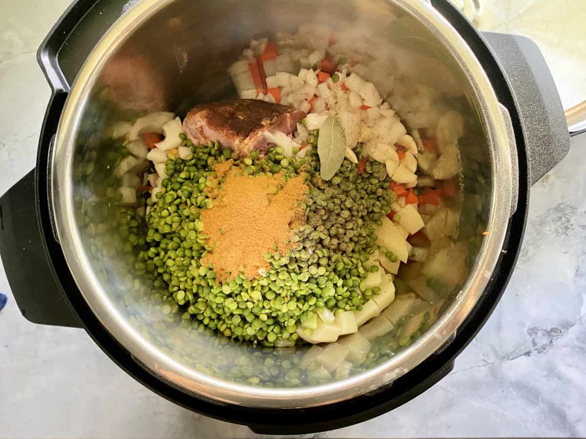 Top view of green split peas, seasoning, ham, onions, carrots, and bay leaf in an Instant Pot.