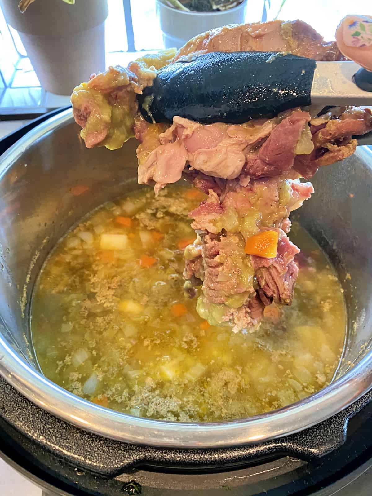 Black tongs holding a cooked ham bone over a pot of split pea soup in an Instant Pot.