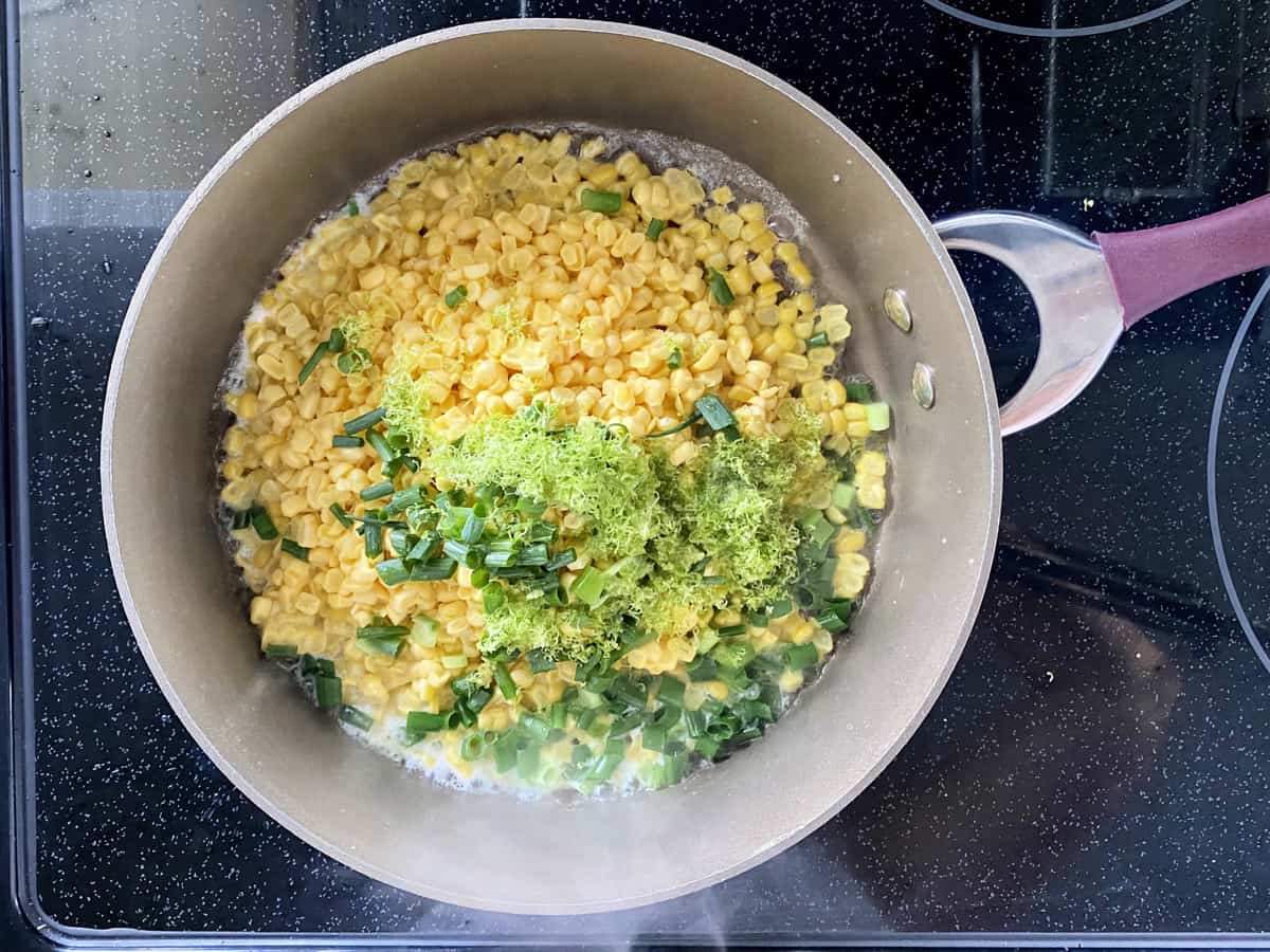 Top view of corn kernels, green onions, and lime zest in a brown skillet on a stovetop.