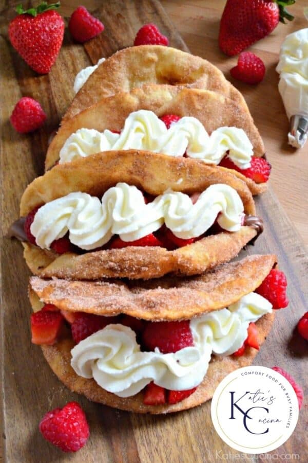 Top view of four dessert tacos topped with strawberries and whipped cream with logo on right corner.