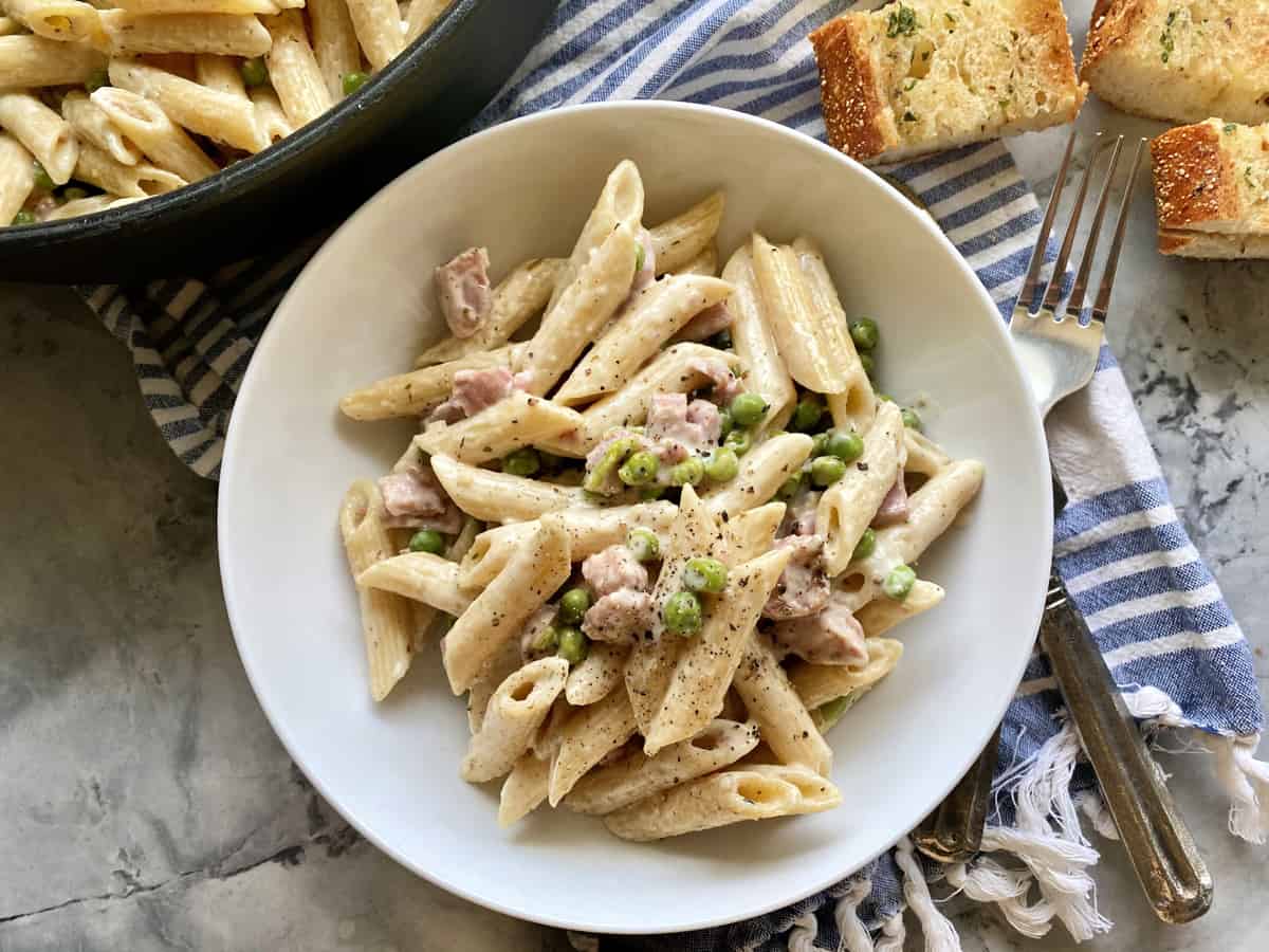 Top view of a white bowl filled with creamy pasta, ham, and peas with a fork and bread next to the plate.