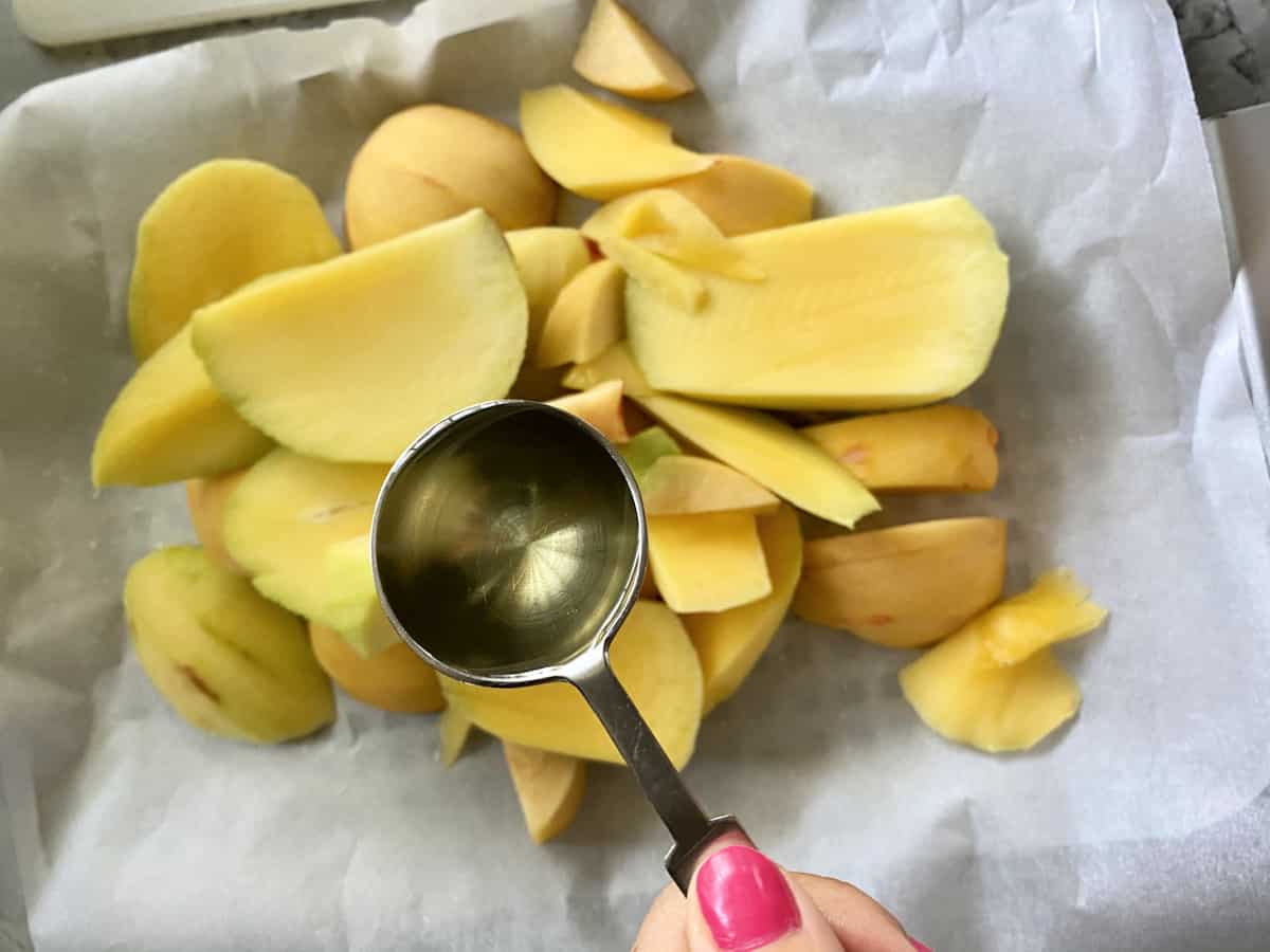 Peeled mangoes and peaches chopped on a parchment line baking sheet with a female hand holding a tablespoon of Mazola® Corn Oil over top.