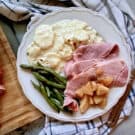 Top view of a white plate on a plaid cloth filled with ham, apples, green beans, and potatoes.