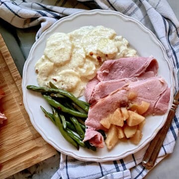 Top view of a white plate on a plaid cloth filled with ham, apples, green beans, and potatoes.