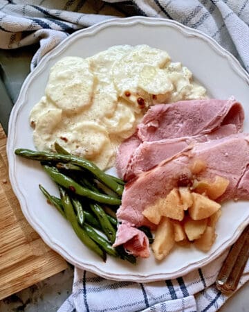 Top view of a white plate on a plaid cloth filled with ham, apples, green beans, and potatoes.