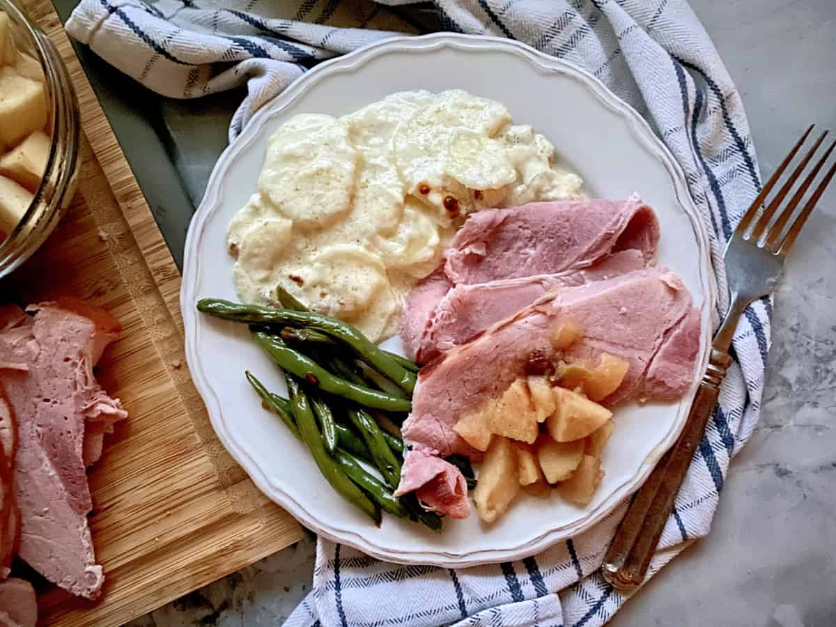 Top view of a white plate on a plaid cloth filled with ham, apples, green beans, and potatoes.