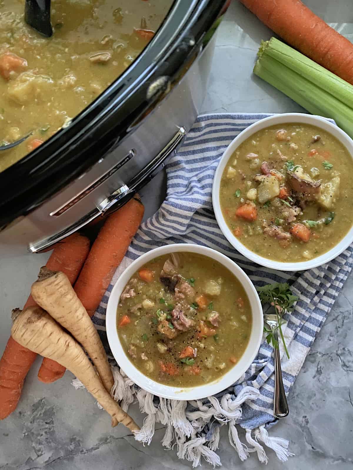 Top view of a slow cooker and two white bowls filled with split pea soup with fresh vegetables on the side.